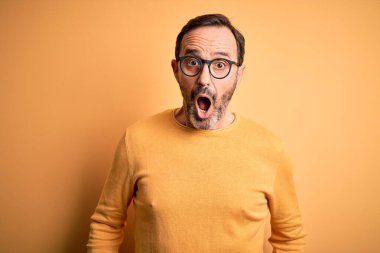 Orta yaşlı, beyaz bir adam, günlük süveter ve gözlük takıyor, sarı arka planda korku ve şaşkınlık içinde, şaşkınlık içinde, korku ve heyecan dolu bir yüz ifadesi var..