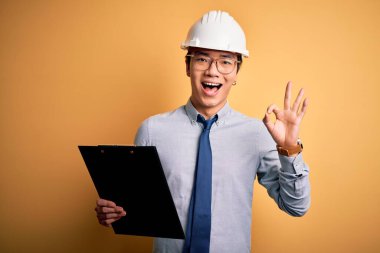 Güvenlik kaskı takan yakışıklı Çinli mühendis elinde pano tutuyor. Parmaklarıyla imza atıyor. Mükemmel bir sembol.