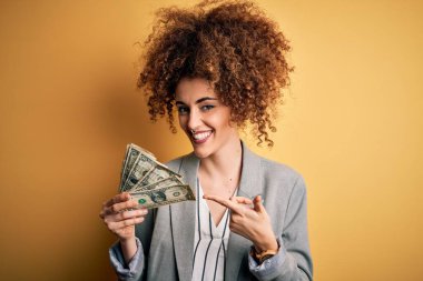 Kıvırcık saçlı ve piercingli genç bir kadın elinde dolarlarla mutlu bir şekilde parmakla işaret ediyor.