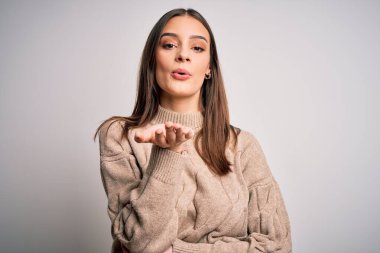 Genç, güzel, esmer, sıradan bir süveter giyen bir kadın beyaz arka planda durup kameraya bakıyor ve havada öpücükler savuruyor. Güzel ve seksi. Aşk ifadesi.