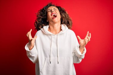 Orta yaşlı, güzel, sportif bir kadın, izole edilmiş kırmızı arka planda beyaz bir kazak giyiyor. Deli gibi bağırıyor, bağırıyor, agresif bir ifade ve kollarla bağırıyor. Hayal kırıklığı konsepti.