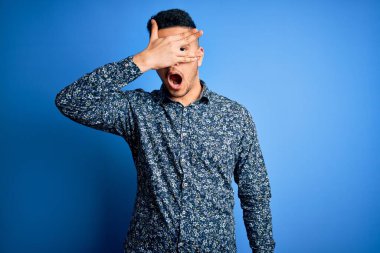 Genç ve yakışıklı bir adam, günlük gömleğini giymiş, mavi arka planda dikiliyor. Şok olmuş bir şekilde yüzünü ve gözlerini kapıyor, utanmış bir ifadeyle parmaklarının arasından bakıyor..
