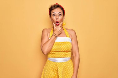 50 'li yaşlarda, sarı arka planda eski moda elbise giyen orta yaşlı bir kadın. İnançsızlığa, şaşkınlığa ve elleri çenesinde olan şaşkınlık ifadesine hayran görünüyor.