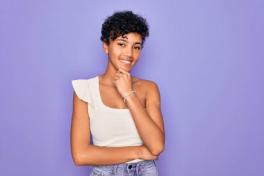 Genç ve güzel Afro-Amerikan kadın mor arka planda rahat bir tişört giyiyor. Kameraya bakıyor. Kollarını kavuşturup, elini çenesine kaldırmış gülümsüyor. Olumlu düşünüyorum..