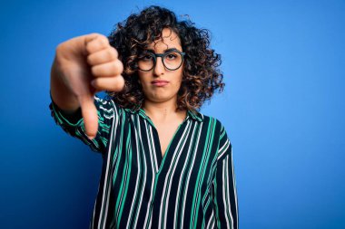 Mavi arka planda çizgili gömlek ve gözlük takan güzel, kıvırcık Arap kadın mutsuz, reddedilmiş ve olumsuz hareketler sergiliyor. Kötü ifade.