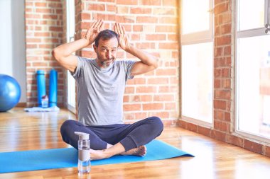 Orta yaşlı yakışıklı sporcu mindere oturmuş spor salonunda yoga egzersizi yapıyor. Elleri kuşkucu ve alaycı görünen el hareketleriyle tavşan kulağı hareketi yapıyor. Paskalya tavşanı kavramı.
