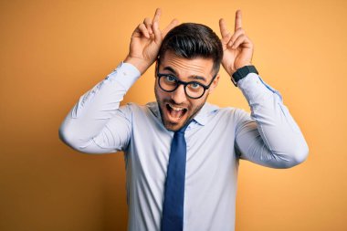 Sarı arka planda duran kravat ve gözlük takan genç yakışıklı iş adamı komik ve deli pozları veriyor parmakları tavşan kulağı gibi, neşeli gülüyor.