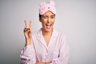 Beyaz arka plan üzerinde pijama ve uyku maskesi takan genç, güzel esmer kadın kameraya gülümseyip zafer işareti yapıyor. İki numara..