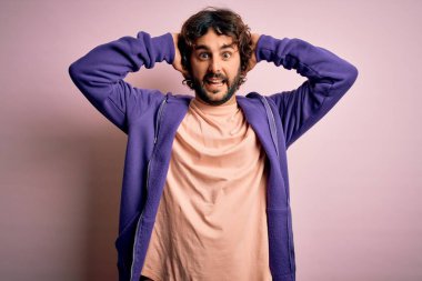 Genç, yakışıklı, sakallı, pembe arka planda sıradan bir kazak giyen, elleri başında, korkmuş ve ağzı açık bir şekilde şok geçirmekten korkan genç bir adam.