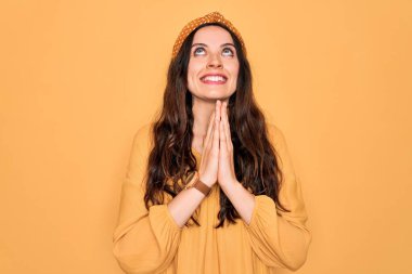 Gündelik tişört ve diyabet giyen genç ve güzel bir kadın sarı arka planda duruyor. Elleriyle yalvarıyor ve dua ediyor. Yüzünde umut ifadesi var. Çok duygusal ve endişeli. Yalvarıyorum..