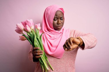 Genç Afro-Amerikan artı boy kadın elinde pembe lale buketiyle Müslüman başörtüsü tutuyor. Saati kontrol ediyor, rahat ve kendinden emin.