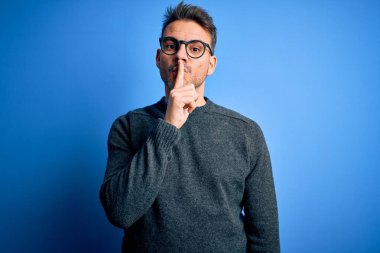 Mavi arka plan üzerinde sıradan süveter ve gözlük takan genç yakışıklı adam sessiz olup dudaklarını öpmek istiyor. Sessizlik ve gizli konsept.