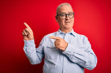 Orta yaşlı, beyaz, sıradan çizgili gömlek ve kırmızı arka planda gözlük takan, gülümseyen ve kameraya bakan, iki eliyle ve parmaklarıyla yüzünü gösteren yakışıklı bir adam..