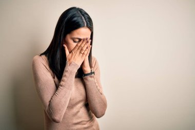 Mavi gözlü genç esmer kadın, izole edilmiş beyaz arka plan üzerine günlük süveter giyiyor ve ağlarken suratını ellerle kapatıyor. Depresyon kavramı.