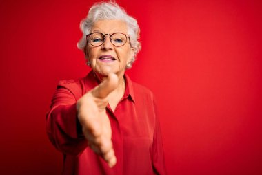 Son sınıf güzeli, gri saçlı, rahat gömlekli ve kırmızı arka planda gözlüklü hoş geldin demek için gülümseyerek tokalaşan bir kadın. Başarılı bir iş..