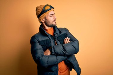 Kar giysileri ve kayak gözlüğü takan sakallı yakışıklı kayakçı kolları çapraz ve kendinden emin bir şekilde yana bakıyor.