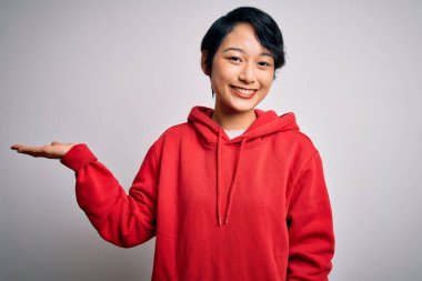 Genç, güzel Asyalı bir kız beyaz arka planda kapüşonlu bir kazak giyiyor neşeli bir şekilde gülümsüyor ve avuç içi kameraya bakıyor..