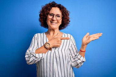 Orta yaşlı, kıvırcık saçlı, sıradan çizgili tişört giyen güzel bir kadın izole arka planda hayrete düşmüş ve kameraya gülümserken eliyle ve parmağıyla işaret ediyor..