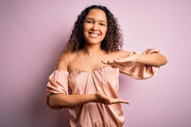 Kıvırcık saçlı, pembe tişörtlü genç bir kadın. Pembe arka planda duruyor. Ellerinde büyük ve büyük beden işareti, ölçü işareti var. Kameraya bakıp gülümsüyor. Ölçüm konsepti.