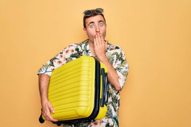 Tatilde mavi gözlü yakışıklı bir turist, sarı arka planda bavulu tutarken ağzını eliyle kapatıyor, şok geçiriyor ve hata yapmaktan korkuyor. Şaşırtıcı ifade.