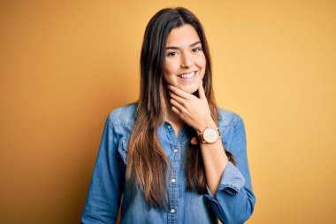 İzole edilmiş sarı arka plan üzerinde duran sıradan bir kot gömlek giyen genç ve güzel bir kız kameraya kollarını kavuşturmuş ve çenesini kaldırmış şekilde gülümsüyor. Olumlu düşünüyorum..