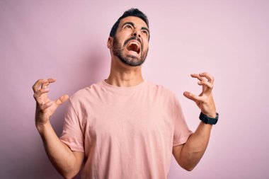 Genç, sakallı, sıradan tişörtlü, pembe arka planda duran çılgın, sinirli, sinirli, agresif ifadeli ve kollarını kaldırmış genç bir adam. Hayal kırıklığı konsepti.