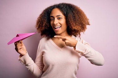 Afro saçlı, pembe arka planda kağıt uçakla gezen Afro Amerikalı genç bir kadın. Elleri ve parmaklarıyla işaret ederek çok mutlu.