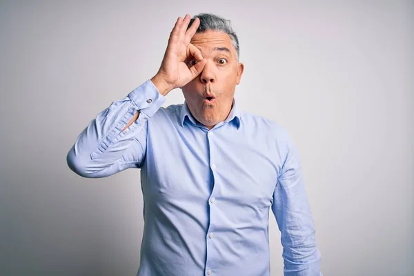 Middle age handsome grey-haired business man wearing elegant shirt over white background doing ok gesture shocked with surprised face, eye looking through fingers. Unbelieving expression.