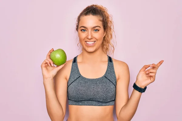 年轻美丽的金发女运动员 蓝眼睛 手里拿着健康的绿色苹果果实 手指手画脚地指向旁边 非常快乐 — 图库照片