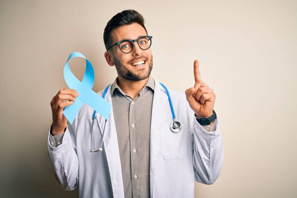 Молодой профессиональный врач, держа в руках голубую ленточку с толстой кишкой на изолированном фоне, удивленный идеей или вопросом, указывая пальцем со счастливым лицом, номер один
