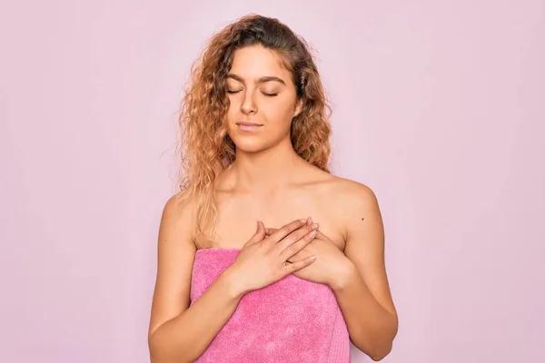 Beautiful blonde woman with blue eyes wearing towel shower after bath over pink background smiling with hands on chest with closed eyes and grateful gesture on face. Health concept.