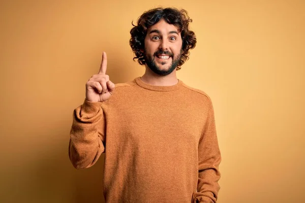 成功したアイデアで指を指して黄色の背景の上に立ってカジュアルなセーターを着てひげを持つ若いハンサムな男 興奮し 第一番 — ストック写真