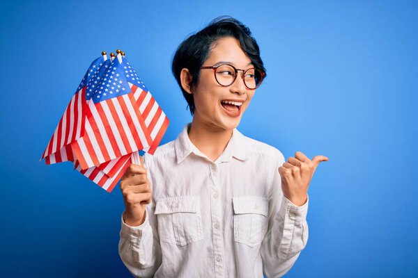 Young beautiful asian patriotic girl holding united states flags celebrating independence day pointing and showing with thumb up to the side with happy face smiling