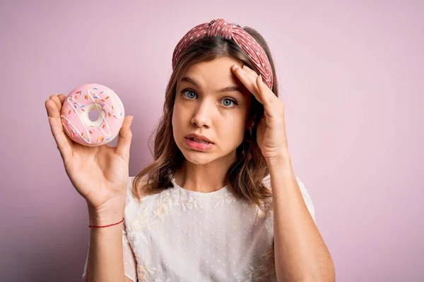若いブロンドの女の子は ピンクの孤立した背景に手で頭の上に強調し 恥と驚きの顔 怒っているとフラストレーションでショックを受けて甘いパン屋さんの砂糖ドーナツを食べている 恐怖と過ちへの動揺 — ストック写真