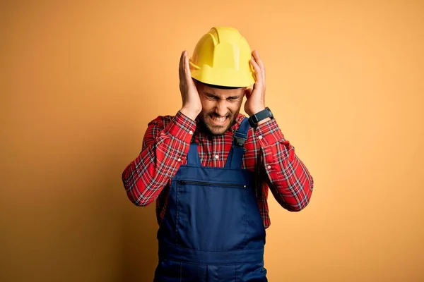 年轻的建筑工人身穿建筑制服 头戴安全帽 头戴黄色的隔离背景 因疼痛和偏头痛而感到绝望和压力 手放在头上 — 图库照片