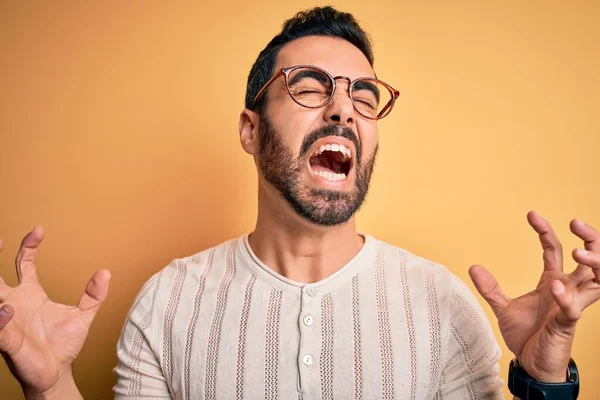 黄色の背景にカジュアルなTシャツと眼鏡を身に着けて髭を生やした若いハンサムな男は狂気と怒りの叫びと積極的な表現と腕を上げて叫んでいます 欲求不満の概念 — ストック写真