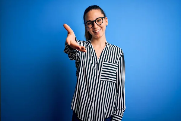 Beautiful blonde woman with blue eyes wearing striped shirt and glasses over blue background smiling friendly offering handshake as greeting and welcoming. Successful business.