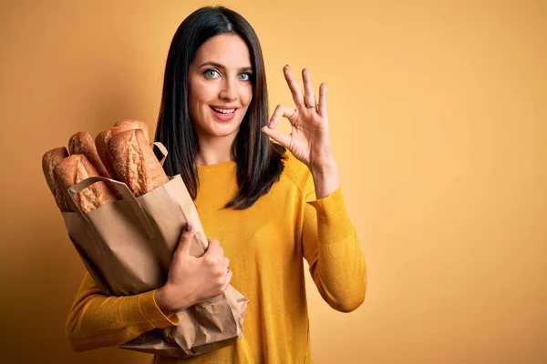 一个蓝眼睛的年轻女人拿着纸袋 上面夹着面包 背景是孤立的 黄色的 手拿着手签 是个很好的符号 — 图库照片
