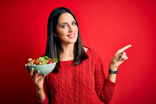 年轻的黑发女人 蓝眼睛 吃着健康的绿色沙拉 而不是孤立的红色背景 她非常高兴地将手和手指指向旁边 — 图库照片