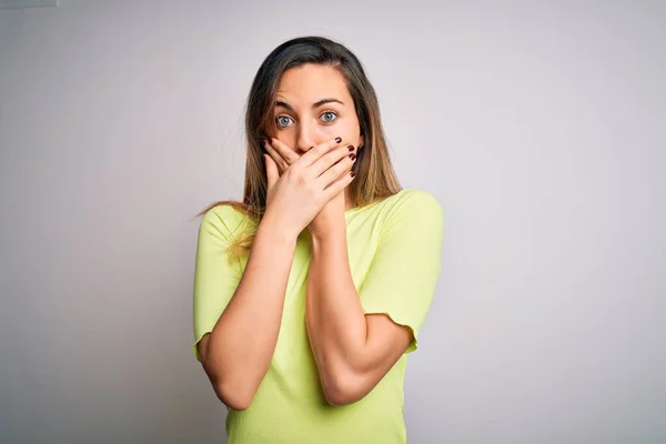 Beautiful blonde woman with blue eyes wearing green casual t-shirt over white background shocked covering mouth with hands for mistake. Secret concept.