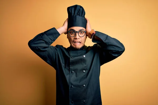 Young brazilian chef man wearing cooker uniform and hat over isolated yellow background Crazy and scared with hands on head, afraid and surprised of shock with open mouth