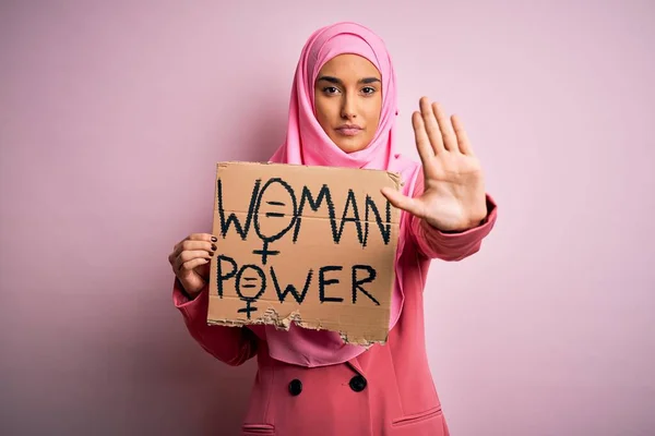 Young activist arab woman wearing pink muslim hijab holding banner with power message with open hand doing stop sign with serious and confident expression, defense gesture