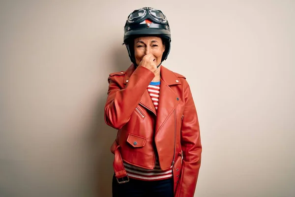 中年のオートバイの女性が白い背景にバイクのヘルメットとジャケットを着て何か臭いと嫌な 耐え難い匂いを嗅ぎ 鼻の上に指で息を保持する 匂いが悪い — ストック写真