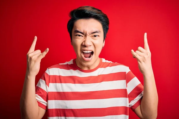 若いハンサムな中国人男性が手を上げて岩のシンボルをやって狂気の表情で叫んで赤い背景の上に立ってカジュアルな縞模様のTシャツを着て 音楽スター 重い音楽の概念 — ストック写真