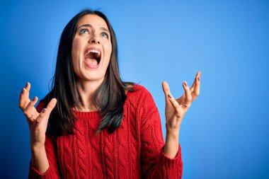 Mavi gözlü genç esmer kadın izole edilmiş arka planda kırmızı bir süveter giyiyor deli deli deli bağırıyor ve agresif bir ifadeyle ve kollarıyla bağırıyor. Hayal kırıklığı konsepti.