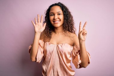 Kıvırcık saçlı, rahat tişörtlü, pembe arka planda duran ve yedinci parmaklarını göstererek gülümseyen, kendinden emin ve mutlu bir kadın..