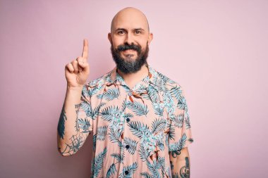 Sakallı ve dövmeli yakışıklı, pembe arka planda sıradan bir çiçek gömleği giyen ve gülümseyen, kendinden emin ve mutlu bir şekilde bir numarayı gösteren kel bir adam..