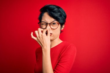 Genç, güzel Asyalı bir kız. Günlük tişört ve gözlük takıyor. Kırmızı arka planda, pis ve iğrenç bir koku var. Kötü kokuyor.