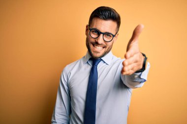 Sarı arka planda duran kravat ve gözlük takan genç yakışıklı işadamı dostça bir selamlaşma ve selamlama olarak el sıkışma sunuyor. Başarılı bir iş..