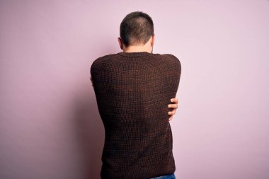 Sakallı ve mavi gözlü genç bir adam pembe arka planda sıradan bir süveter giyiyor. Kendine sarılıyor, mutlu ve pozitif bakıyor. Sevgi ve özgüven.